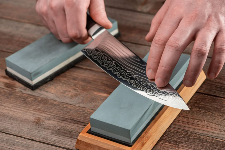 Sharpening a Damascus steel chefs knife on whetstones in a rustic kitchen scene.