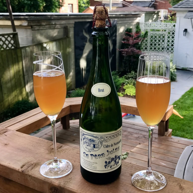Outdoor celebration with Le Pere Jules cidre de Normandie, flutes, and lush garden background.