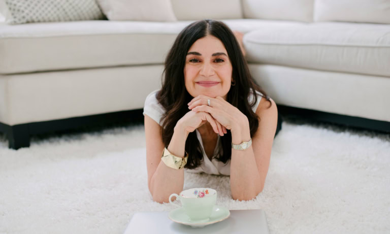 Elizabeth Wanless relaxes on a cozy rug with a smile, tea cup, and laptop nearby.