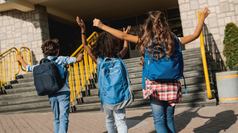 Children going to school joyfully, backpacks on, walking down stairs in bright sunlight.