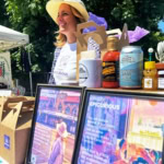 Vibrant outdoor scene with a smiling woman at a farmers market stand showcasing local foods and products.