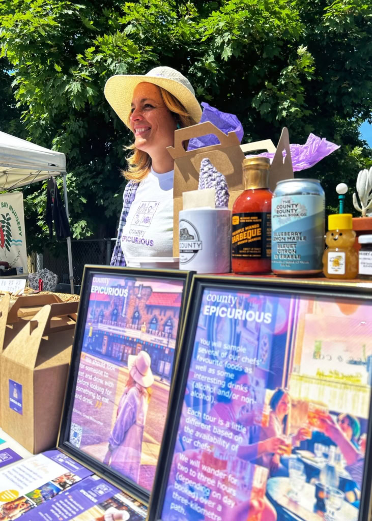 Vibrant outdoor scene with a smiling woman at a farmers market stand showcasing local foods and products.