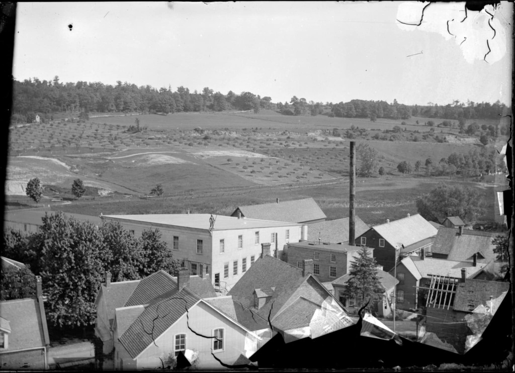 Black and white photo of a small town nestled in rolling hills, featuring houses, trees, and open farmland in the background.