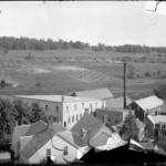Black and white photo of a small town nestled in rolling hills, featuring houses, trees, and open farmland in the background.