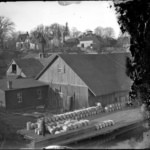 A historical black-and-white image featuring rustic wooden barns, a lumberyard, and a serene rural village backdrop, ideal for showcasing traditional architecture, rural life, and vintage scenes.