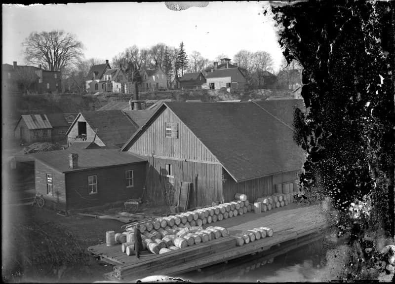 A historical black-and-white image featuring rustic wooden barns, a lumberyard, and a serene rural village backdrop, ideal for showcasing traditional architecture, rural life, and vintage scenes.