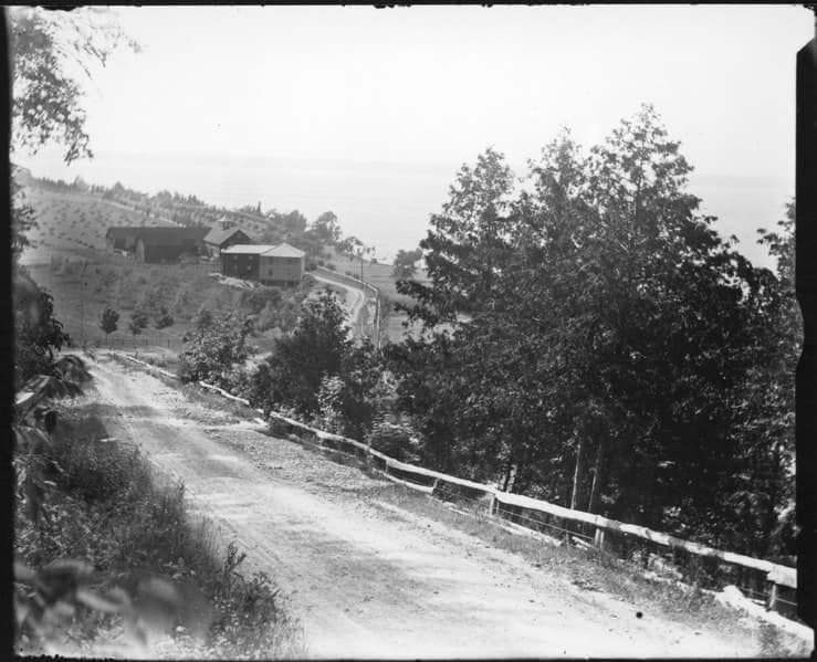 Black and white photo of a peaceful rural scene with dirt road, trees, and hillside houses.