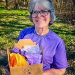 Elegant woman smiling outdoors, holding a "County Epicurious" food box filled with colorful tissue paper.
