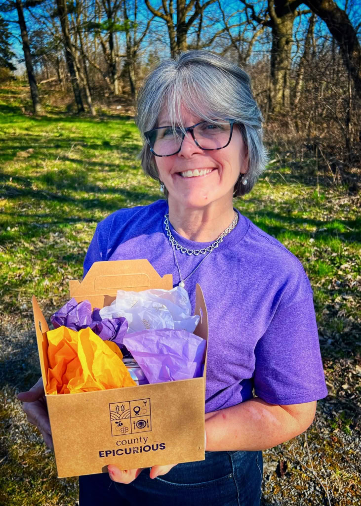 Elegant woman smiling outdoors, holding a "County Epicurious" food box filled with colorful tissue paper.