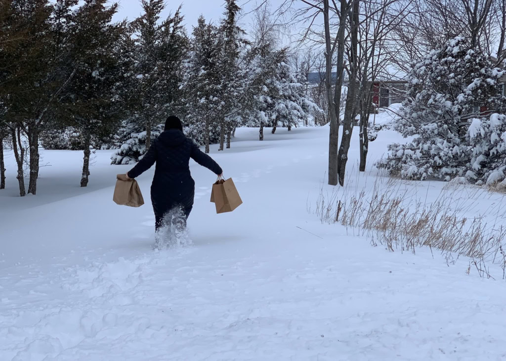 A person in a dark coat and hat walks through snowy terrain, carrying paper grocery bags, during winter.