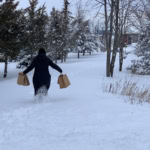 A person in a dark coat and hat walks through snowy terrain, carrying paper grocery bags, during winter.