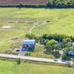 Aerial view of rural farmhouse with organic vegetable garden, lush greenery, and country road integration for farm-to-table dining experience.