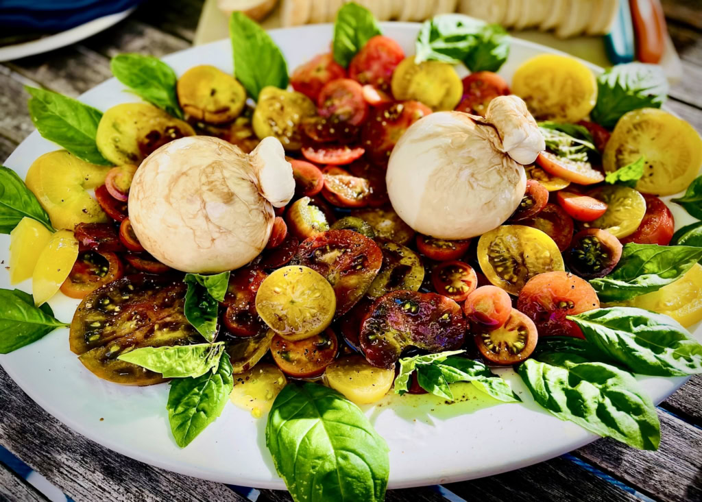 Colorful heirloom tomatoes, fresh basil leaves, and mushrooms on a white platter, perfect for gourmet catering.