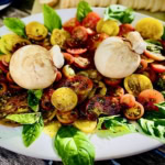 Colorful heirloom tomatoes, fresh basil leaves, and mushrooms on a white platter, perfect for gourmet catering.
