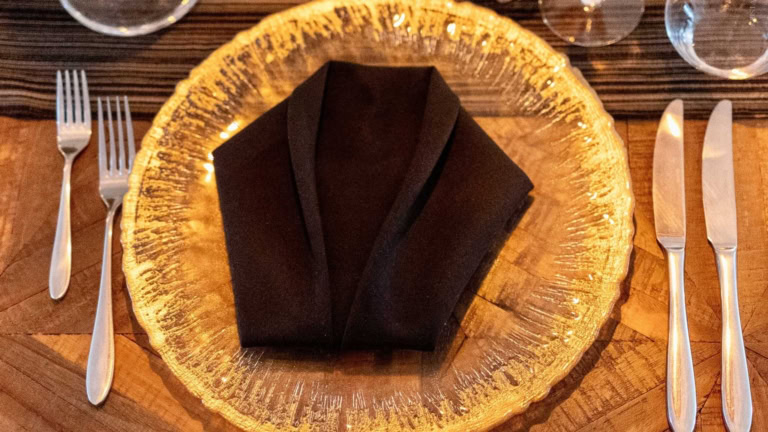 Elegant dinner table with gold plate, black napkin, silver flatware in intimate setting.