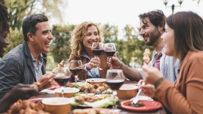 Friends celebrating outdoor dinner with wine and diverse food on a sunny day.