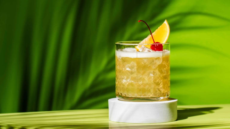 Golden whiskey sour cocktail with ice, cherry, and lemon, set against a tropical background.