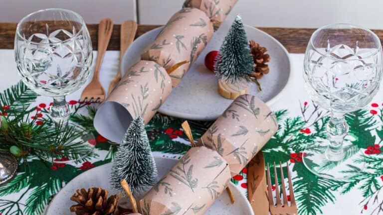 Elegant holiday dinner table with wrapped napkins, pinecones, mini Christmas trees, and glassware.