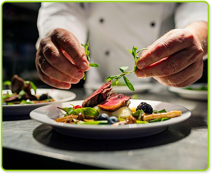 Chef plating gourmet dish with herbs in dimly lit professional kitchen.