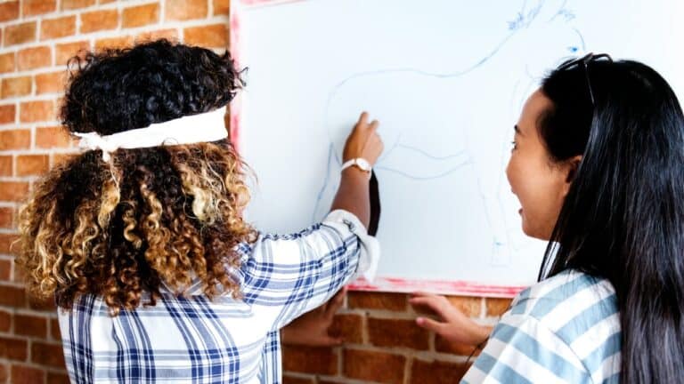 Two women playing a fun party game at a gathering, drawing on a whiteboard.