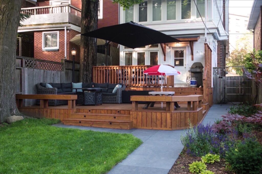 Stylish wooden deck with seating, shade sail, striped umbrella, and lush greenery.