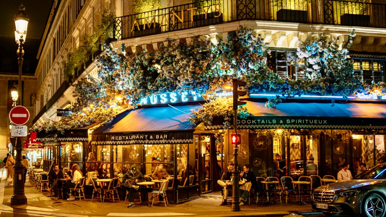 Charming nighttime street scene with lively restaurant, diners, greenery, and Parisian atmosphere.