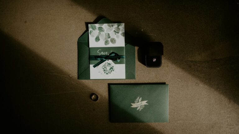 Elegant wedding invitation flat lay with green envelope, floral card, gift box, and engagement ring.