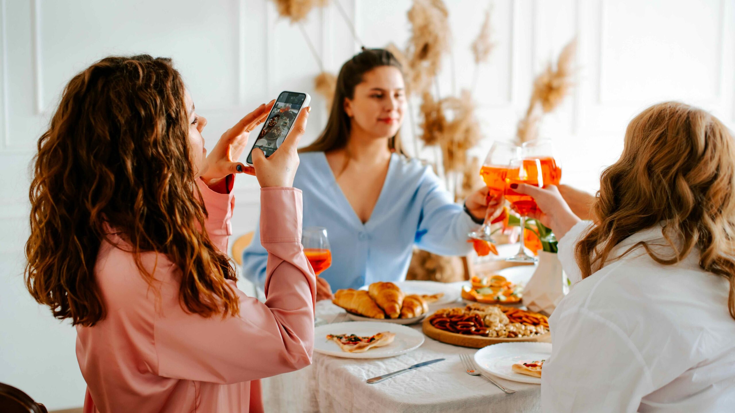 Women friends toasting at brunch table, photographing dinner party, cozy indoor celebration with pastries.