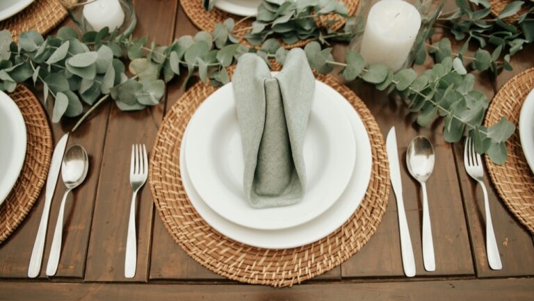 Elegant rustic dining table with woven placemats, white plates, green napkins, and eucalyptus centerpiece.
