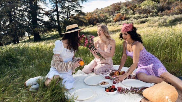 Three women enjoying a boho picnic in a lush green field with flowers and snacks.