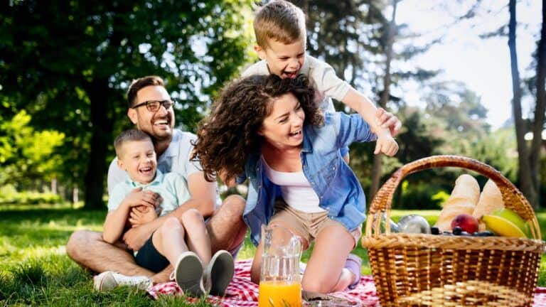 Family enjoying a picnic outdoors with a basket of food and drinks.