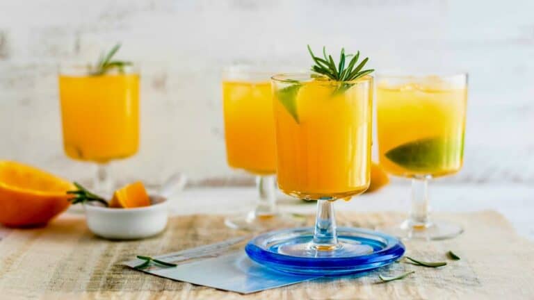 Bright mango cocktails garnished with lime and rosemary on a wooden table.