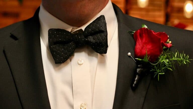 Elegant groom in black tuxedo with red rose boutonniere and black bow tie at wedding reception.