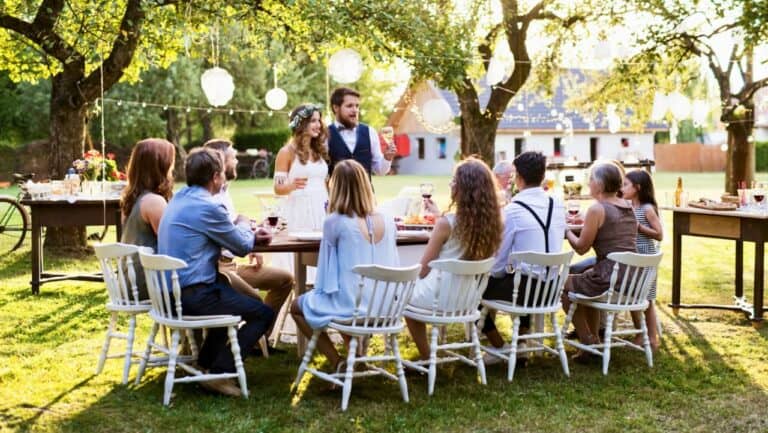 A lush outdoor wedding reception with guests seated around a long table under trees.