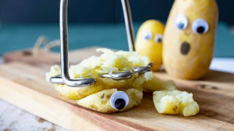 Fun potato with googly eyes being mashed with a potato masher on a wooden board.