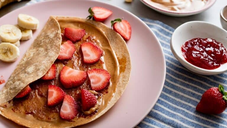 Delicious strawberry and banana crepe with fruit toppings and jam on a pink plate.