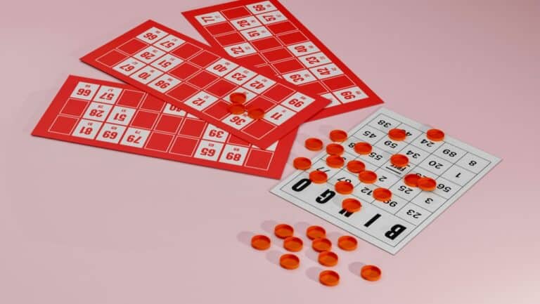 Red and white bingo cards with orange and black bingo chips scattered on a light pink background.
