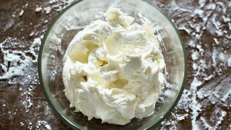 Cream cheese in a glass bowl on a wooden surface with flour scattered around.
