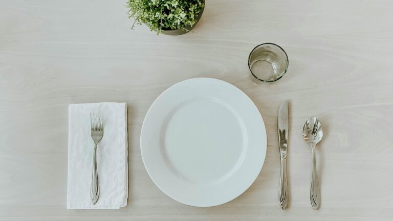 A clean, empty white plate with silverware and a small potted plant on a light wooden table.