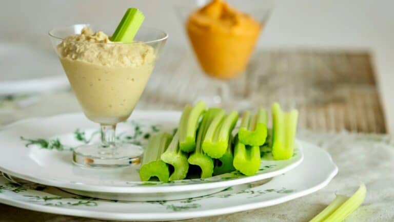Creamy hummus served with fresh celery sticks on a white plate, with a blurred background of additio.