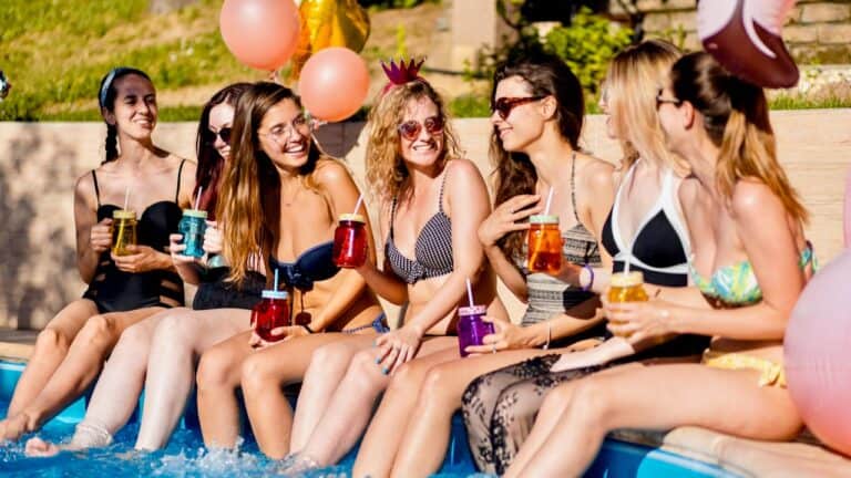 Group of women enjoying drinks at a poolside party with balloons and sunny weather.