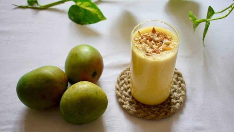 Fresh green mangoes next to a creamy mango smoothie garnished with shredded coconut on a white surfa.