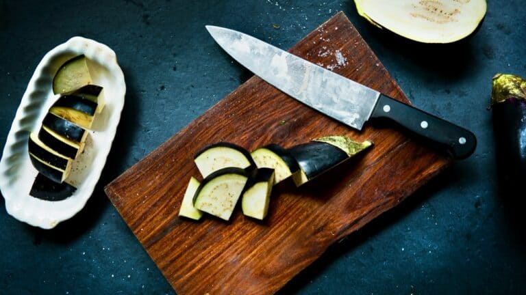 Sliced eggplant on a wooden cutting board with a chef's knife and fresh eggplant in the background.