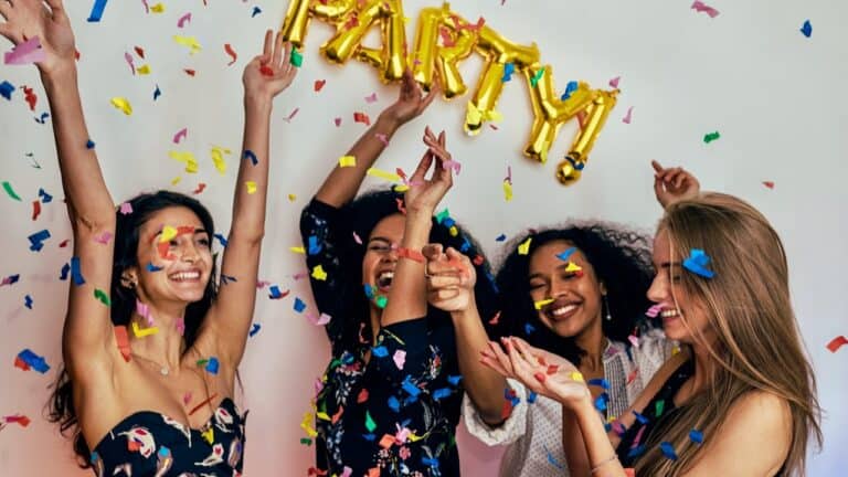 Brightly dressed women celebrating with confetti and gold "PARTY!" balloons at a festive gathering.