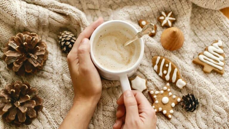 A person holding a mug of hot beverage surrounded by Christmas cookies and pinecones.
