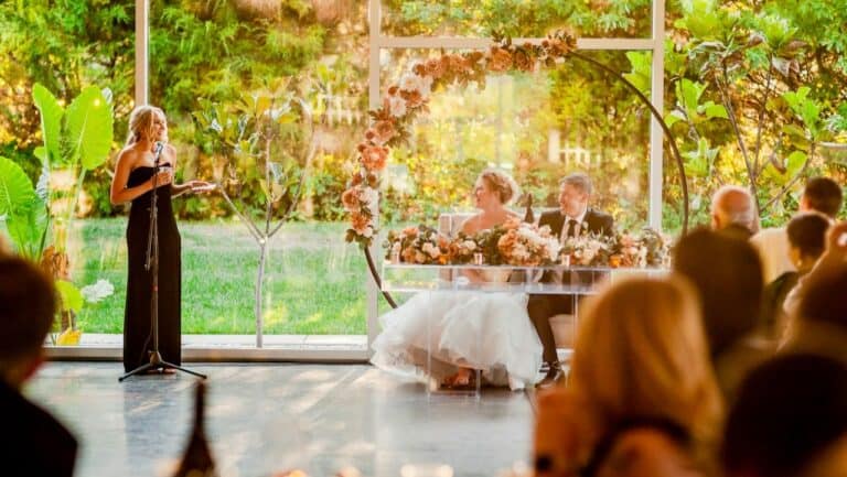 Wedding reception with bride, groom, and guests in a glass-walled venue surrounded by greenery.