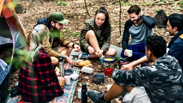 Group of friends enjoying a camping meal outdoors in a forest setting.