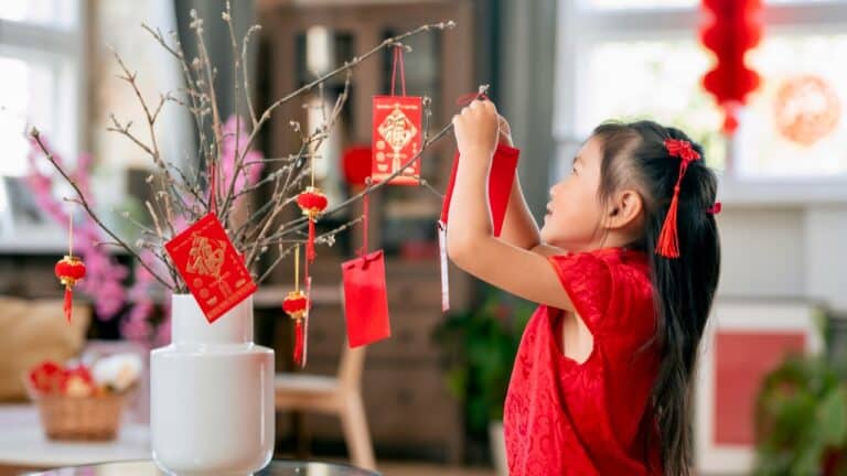 Young girl in red dress decorating a Lunar New Year tree with red envelopes and ornaments.