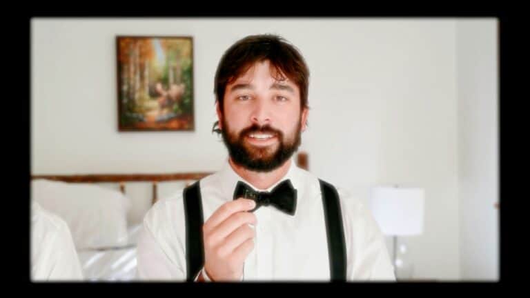 Man with a beard in a tuxedo holding a bow tie in a bedroom setting.